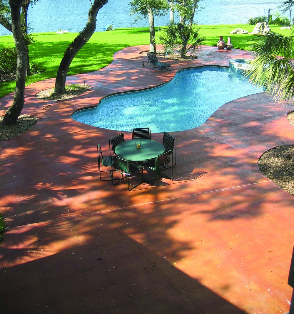 Huge concrete deck surrounding a kidney shaped swimming pool next to a lake.