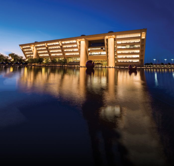 A large building with bright windows and columns made of concrete reflecting off of a nearby body of water.