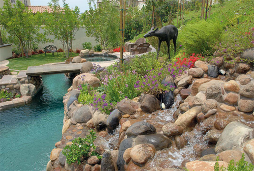 A multilevel waterfall splits into several spillways as it cascades toward a pool. The cast concrete bridge was stamped to look like it was made from wood planks. Colorful plants were used to soften the hardscaped areas.